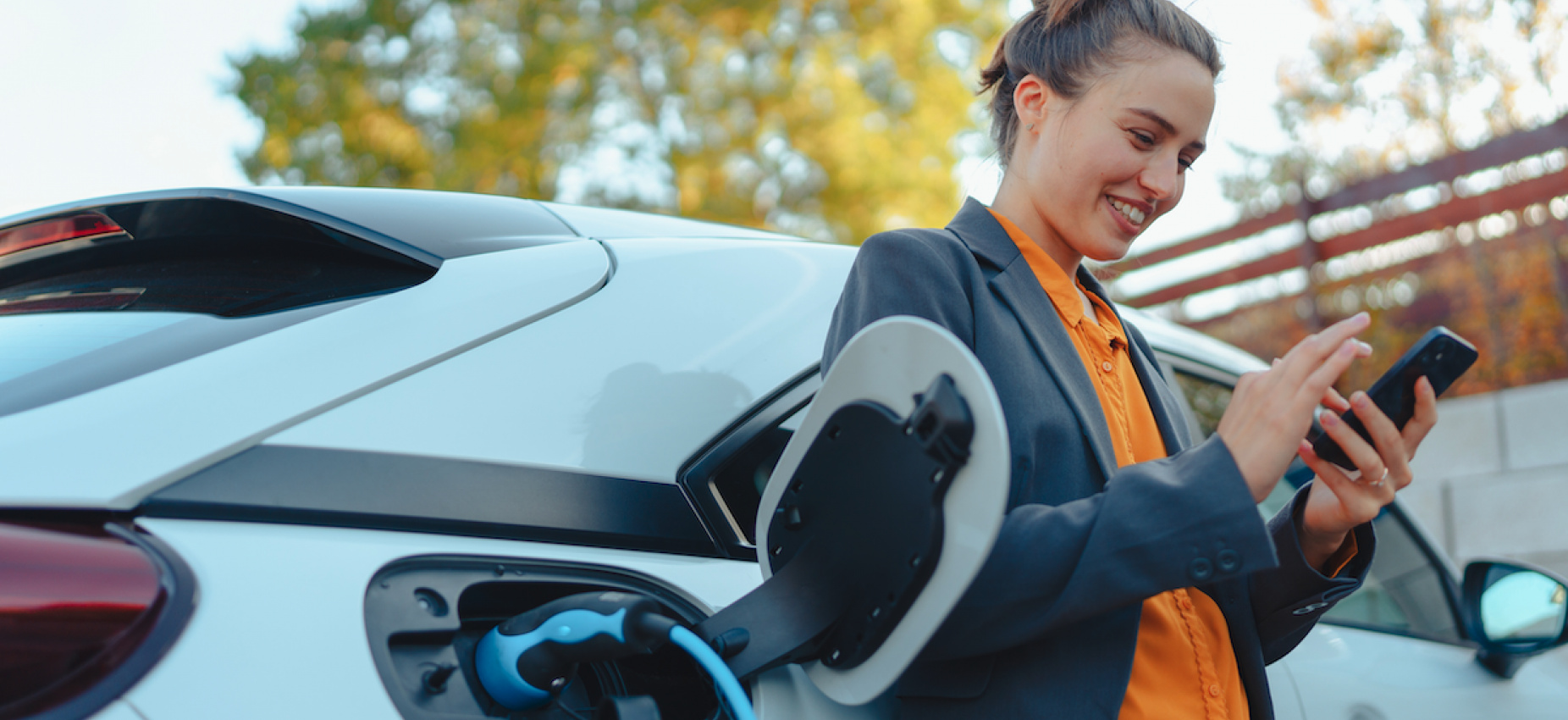 Smiling Woman dressed in work wear charging her EV while using her mobile phone