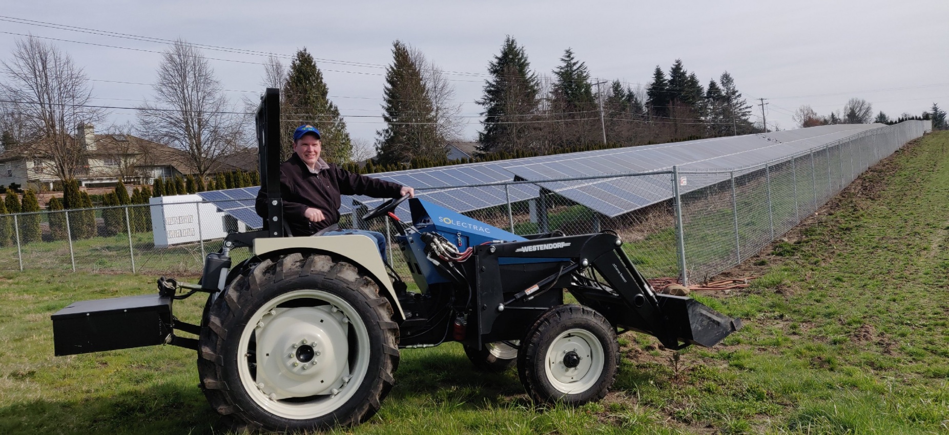 Man atop an electric tractor on a farm with solar panels in the background