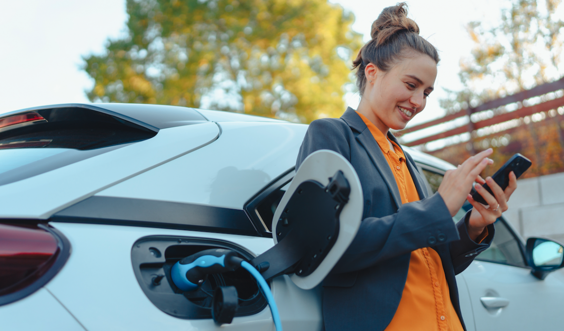 Woman in business attire smiles while leaning on her charging EV with phone in hand